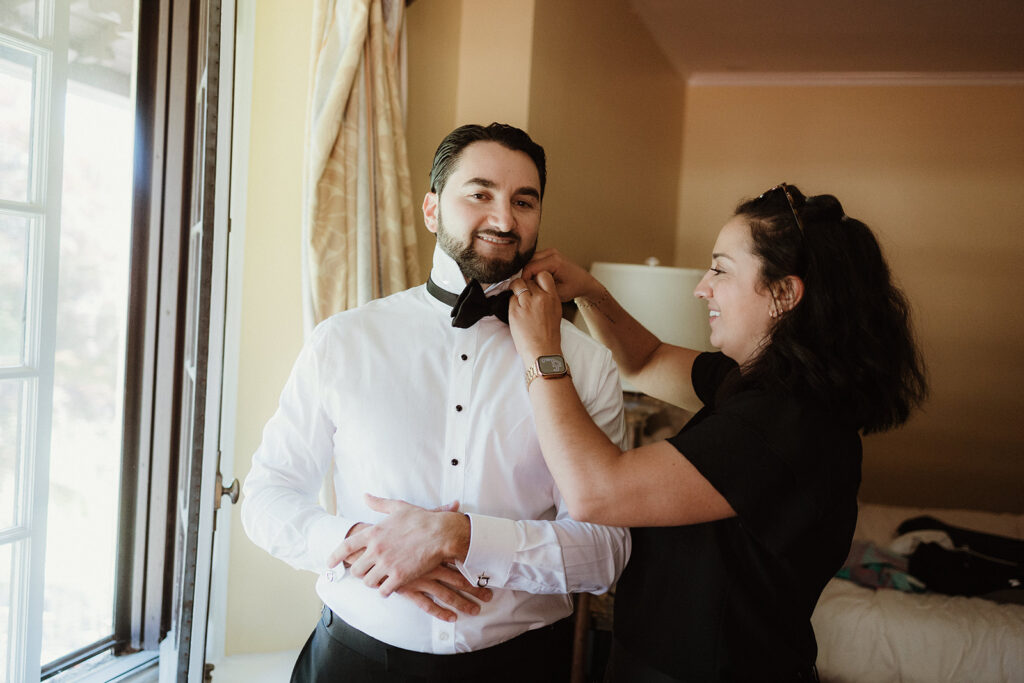 Kelly jeanmaire, Wedding planner at The Main Event by Kelly, putting a bow tie on a groom at the Rushmore Estate in Highland Mills new york. Very high end wedding venue in the Hudson Valley.